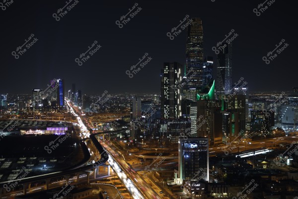 A nighttime image of Riyadh in Saudi Arabia, showing a cluster of illuminated skyscrapers in the city center. The streets are lit with bright lights with visible traffic, and the Kingdom Tower stands out with its distinctive colors. The urban lighting creates a vibrant atmosphere in the city.