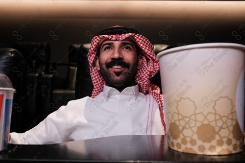 شاب سعودي يرتدي الزي السعودي التقليدي يستمتع بمشاهدة الفيلم في صالة السينما ويظهر أمامه الفشار