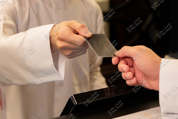 Two individuals in white clothing exchanging a rectangular card over a counter. The image shows a hand in traditional attire receiving the card from another person, in a setting that may be a reception desk or a store.