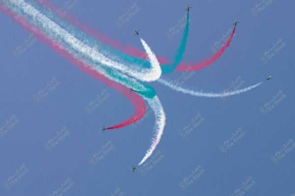An image showing a group of airplanes flying in a clear sky, leaving behind a trail of smoke in various colors including white, blue, and red. The aircraft are performing stunning aerobatic maneuvers, creating artistic paths with the colored smoke.