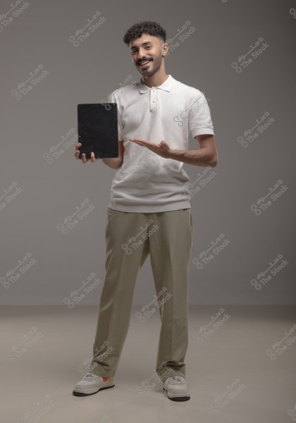 A man stands smiling in a studio, wearing a white polo shirt, beige trousers, and sneakers. He holds a tablet in his right hand and gestures towards it with his left hand. The background is a plain gray.