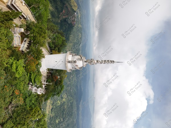 The image shows a tall white tower adorned with spiral details at the top, surrounded by dense green trees and mountainous landscapes in the background. The sky is cloudy, adding a sense of towering elevation to the scene.