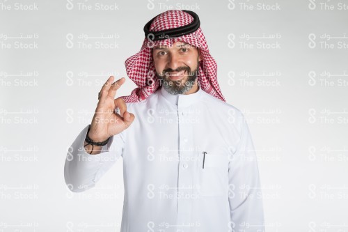 بورتريه رجل سعودي يقف على خلفية بيضاء، يرتدي الثوب، ينظر الى الكاميرا وهو مبتسم ويقوم بعمل حركات بيديه، تعبير على اتمام شيء او الاتفاق مع شيء