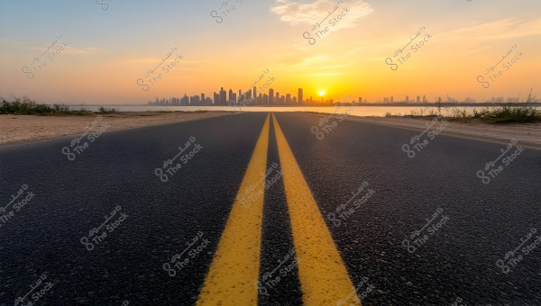 A long road stretches towards the skyline of a modern city during sunset. The image shows yellow lines in the middle of the road, with the city skyline featuring tall buildings in the background, as the sun casts warm colors across the horizon. There is sand and sparse vegetation on both sides of the road.