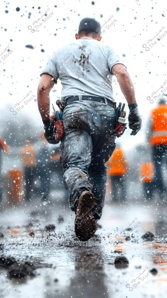 A person wearing a gray shirt and jeans is walking on a muddy ground while water and mud droplets scatter around. The person has tools in their pockets, and the ground is wet. In the background, several workers are visible wearing orange reflective vests.