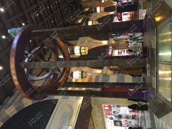 Interior view of a shopping mall showcasing traditional Middle Eastern architectural design. The image includes ornate columns and a large metallic sphere structure inspired by ancient astronomical tools. The lighting is warm with hanging chandeliers, and there are shops in the background displaying illuminated signs.