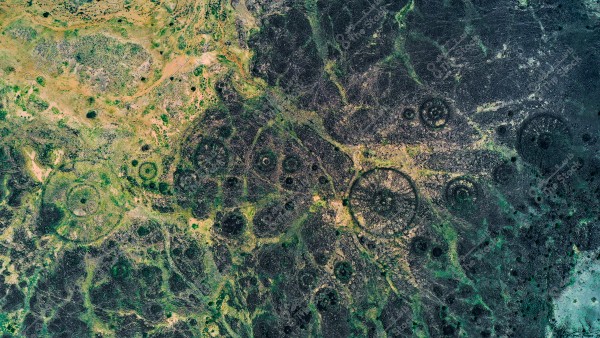 صورة جوية في وادي حلي و مذيلات حجرية تتضمن تشكيلات حجرية دائرية معقدة. تظهر الأراضي بألوان متنوعة من الأخضر إلى البني الداكن, مع انتشار التشكيلات الصخرية التي تشبه دوائر أو عجلات في مختلف الأنحاء. الطبيعة المحيطة تبدو مقفرة بشكل كبير مع بعض النباتات الصغيرة المنتشرة.