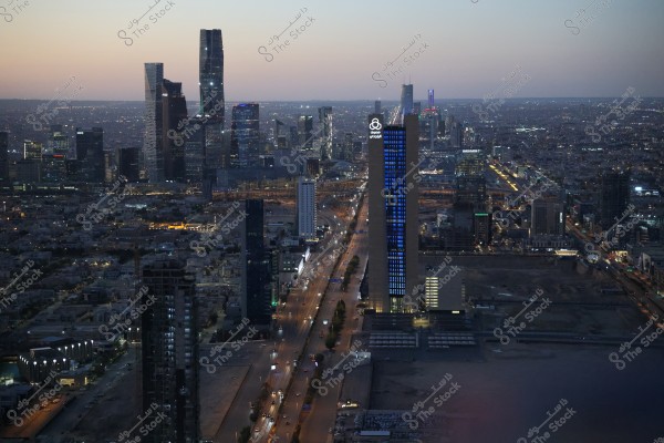 صورة ليلية لمدينة الرياض في المملكة العربية السعودية, تُظهر أفق المدينة المضاء بالكثير من ناطحات السحاب الحديثة, منها برج بارز يحمل شعار فيرجن ميقا ستور مضاء بالأزرق. يظهر الطريق الرئيسي ممتدًا إلى الأمام محاطًا بالمباني الحديثة. السماء تبدو بلون خفيف نتيجة لإضاءة المدينة.