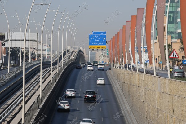 A wide highway stretches in a city that appears to be in Saudi Arabia, with cars traveling in both directions. A blue sign indicates \"Eastern Ring Road\" along with other signs on the highway. The side walls are equipped with vertical metal decorations colored red and orange. On the left side, there is an elevated train track supported by tall white poles.