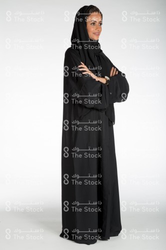 Portrait of a Saudi woman stands on a white background, wears a black hijab and abaya, puts her hands on her arms and smiling