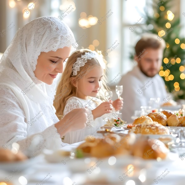 An image of a family celebrating together at a special occasion. A woman wearing a white lace hijab and dress is sitting next to a young girl in a white dress with a matching hairband. On the table in front of them are plates with pastries and various foods. In the background, a man dressed in white is visible, along with part of a tree decorated with illuminated lights, creating a festive and warm atmosphere.