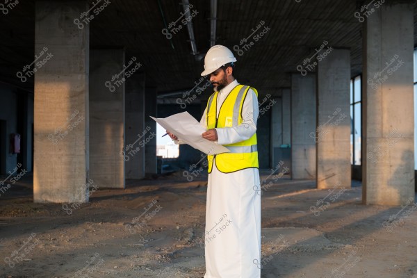 The image shows a man wearing a white thobe and a bright yellow safety vest and gloves, along with a white safety helmet. He is standing inside a building under construction, examining a blueprint or paper in his hand. The surroundings feature concrete pillars and an unfinished dirt floor.