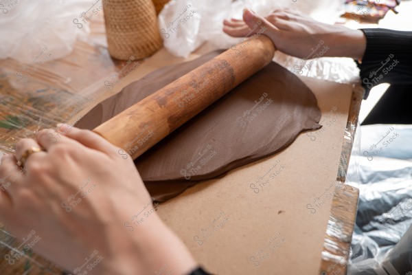 The image shows a person\'s hands using a wooden rolling pin to flatten dark brown dough on a surface covered with clear plastic. In the background, there is a rattan cone and some tools.