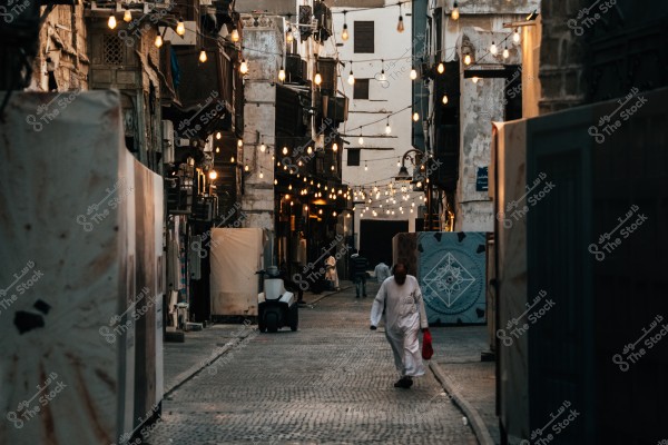 A narrow street in an old city with traditional Arabic architectural buildings. String lights hang along the street, adding a warm atmosphere. A man wearing traditional white attire carries a red bag while walking on the cobblestone pavement. A small scooter is parked beside the road.
