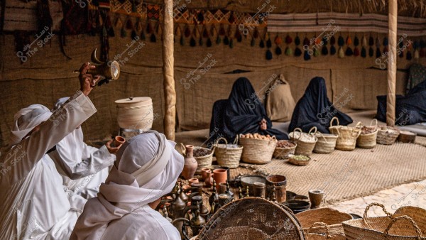 Inside a traditional tent, men dressed in white thobes and shemaghs are gathered. One man is holding up a coffee pot, while others are seated around a table with coffee cups and wicker baskets filled with dates and nuts. In the background, women wearing black abayas are seated next to the baskets. The decor includes fabric walls adorned with traditional designs. The scene suggests a setting from the Arabian Gulf region.