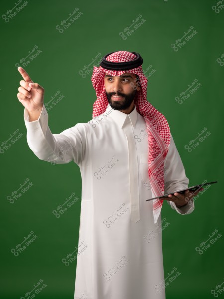 صورة لرجل يرتدي الزي السعودي التقليدي, حيث يظهر مرتدياً الثوب الأبيض والغترة الحمراء والعقال الأسود. الرجل يبتسم ويشير بيده اليمنى بينما يمسك بجهاز لوحي في يده اليسرى. الخلفية خضراء بسيطة.