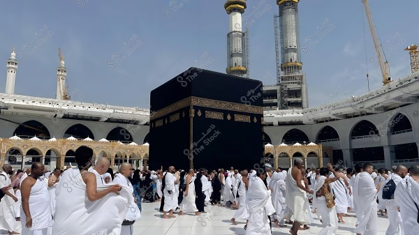 A group of pilgrims performing Tawaf around the Kaaba in the Grand Mosque in Mecca, wearing white Ihram garments, with the mosque\'s minarets in the background.