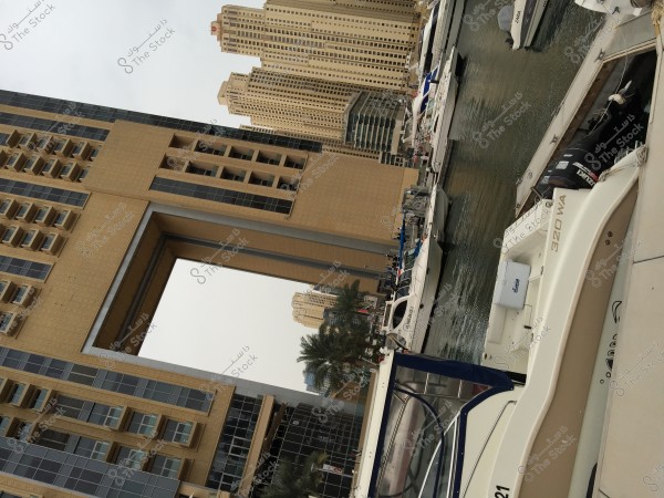 View of a water canal surrounded by tall, modern buildings in an urban area. Boats are moored along the canal, and the buildings feature intricate architectural designs with brown and sandy colors. Palm trees can be seen between the buildings, adding a touch of nature to the scene.