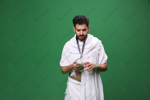 Portrait of a Saudi pilgrim on a green background wearing the Ihram to perform Hajj, carrying a mobile phone and reviewing the green health status