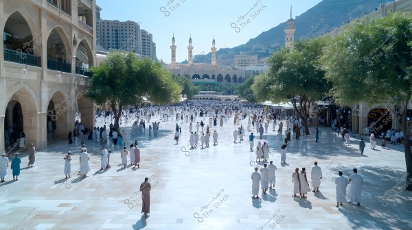 A large courtyard in Medina, showing many people wearing traditional white attire for men in Saudi Arabia. People are walking in the tree-shaded courtyard. In the background, there are large buildings and minarets. The area is surrounded by structures with large arches.