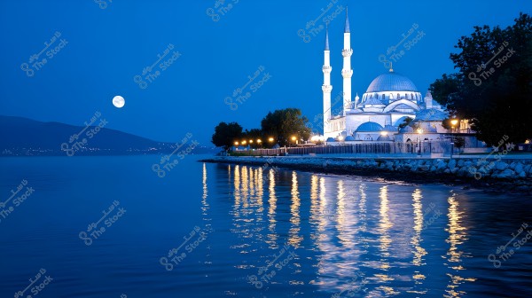 An image of a large mosque illuminated at night by the sea. The full moon is visible in the blue sky, and the mosque\'s lights and lamps reflect on the water\'s surface. Trees and buildings surround the mosque, which features tall minarets and exterior domes.