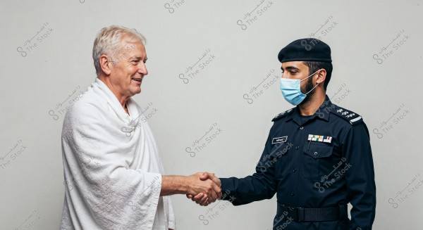 The image shows two men shaking hands. The man on the left is wearing a white Ihram, a garment traditionally worn by Muslims during Hajj and Umrah rituals. The man on the right is in a dark security uniform with a cap, possibly from an Arab country, as indicated by the Arabic name tag. He is wearing a blue mask, suggesting he is following health safety protocols.