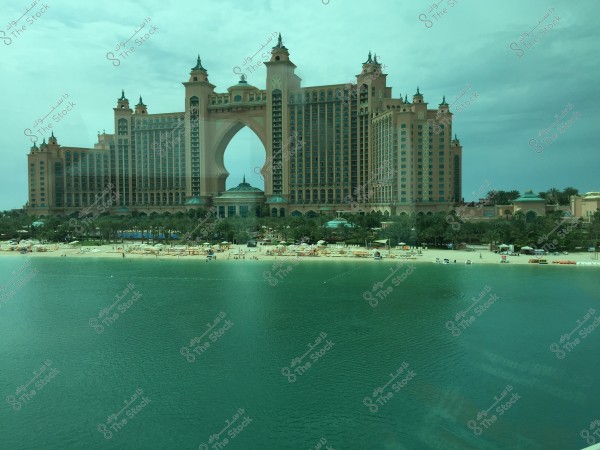 Image of the large Atlantis Hotel with its arched structure located on Palm Jumeirah Island in Dubai, United Arab Emirates. The hotel is surrounded by pools, palm trees, and white sandy beaches, with the calm waters of the gulf in the foreground. The sky is overcast, adding a serene feeling to the overall scene.
