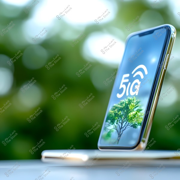 A smartphone positioned on a table, slightly tilted, displaying a \"5G\" logo in white on its screen with an image of a green tree in the background. The image background is blurred with natural green tones, suggesting an outdoor setting.