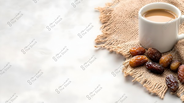 A cup of milk coffee placed on a piece of light brown burlap fabric, surrounded by a variety of dates of different sizes on a blurred background.
