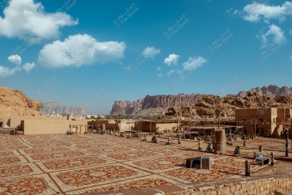 A scenic view of a desert area featuring a large courtyard covered with traditional decorative rugs. Surrounding the courtyard are mud-brick buildings and wooden furniture. Towering rocky mountains form the backdrop, with a clear blue sky and scattered clouds.