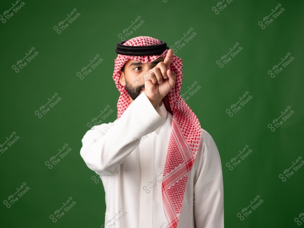 A portrait of a man wearing a traditional white thobe with a red and white checkered headscarf (ghutra) and an agal, standing against a green background and pointing his finger forward.