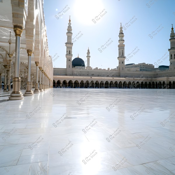 A view of the Prophet\'s Mosque in Medina, Saudi Arabia. The image shows the mosque\'s expansive courtyard made of white marble, with the sun reflecting on the shiny surface. In the background, the mosque\'s green domes and tall minarets are visible under a clear blue sky.