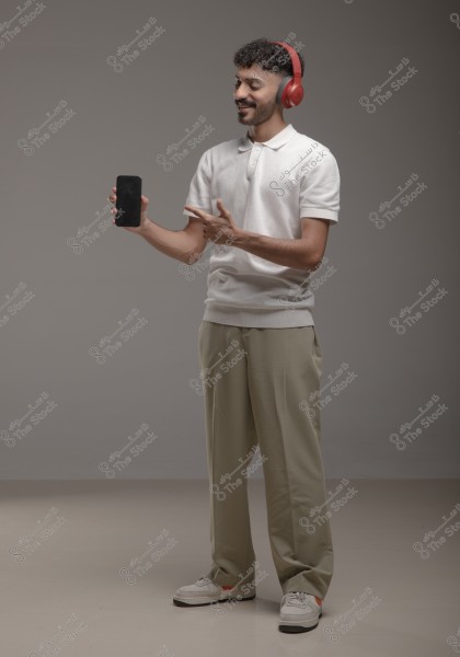A portrait of a person wearing red headphones, holding a smartphone in their left hand, and pointing at it with their right hand. The person is wearing a white shirt and beige pants with white sneakers. The background is a simple grey.