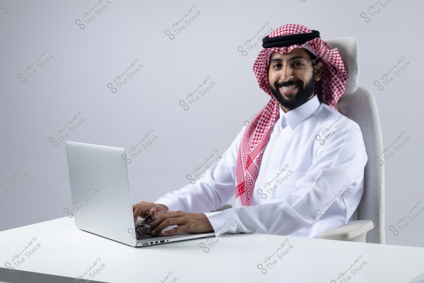 A portrait of a man sitting on a chair wearing traditional Saudi attire, including a white thobe and a red ghutra with a black agal. The man is smiling and working on a silver laptop on a white desk against a neutral gray background.