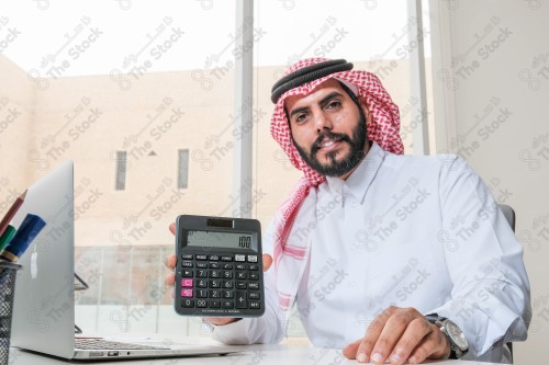 رجل سعودي يرتدي الزي السعودي التقليدي يعمل في مكتب بواجهة زجاجية ويستخدم آلة الحاسبة أثناء العمل