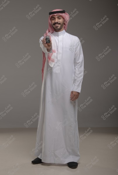 A portrait of a man wearing a white thobe and a traditional red and white headscarf, standing in a studio with a light gray background. The man is smiling and holding a car key in his extended right hand. The attire suggests traditional Saudi style.