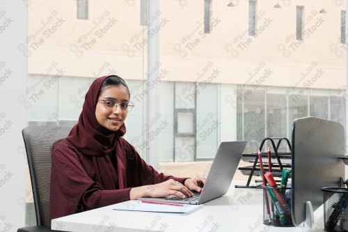 A Saudi woman wearing an abaya works in a glass-fronted office and uses a mobile device to write while taking notes during the day