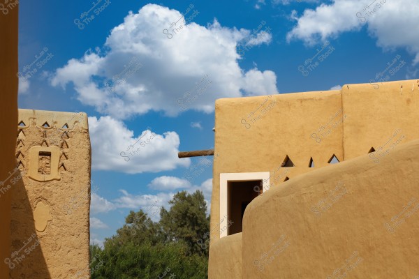 The image depicts a scene of traditional mud architecture under a blue sky with scattered clouds. The walls are decorated with triangular geometric patterns and a small window in the center. In the foreground, broad mud surfaces are visible. The background is adorned with green trees.