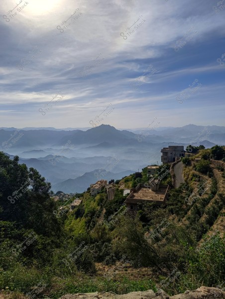 An image showcasing a natural landscape with lush green mountains, creating a breathtaking panoramic view. Several buildings are perched on a hillside, with a narrow road winding around the hills, and distant mountains shrouded in a light mist under a blue sky with wispy clouds.