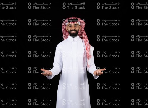 بورتريه رجل سعودي يرتدي الزي الثوب، يقف على خلفية سوداء ويقوم بعمل حركات بيديه لعرض او وصف منتج، رجل يقوم بعمل حركات بيديه وهو ينظر الى الكاميرا