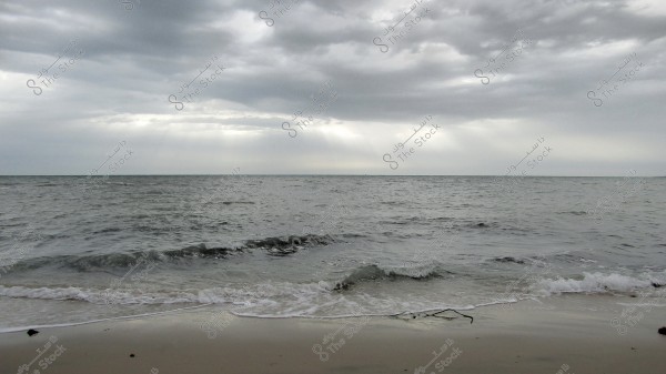 An image of a calm beach where small waves gently hit the sandy shore. The sky is overcast with thick grey clouds, and soft sunlight streams through, reflecting on the surface of the sea.