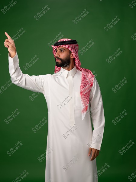 A portrait of a man wearing a white thobe and a red checkered headscarf with a black agal, standing and pointing upward with one hand. The background is green.