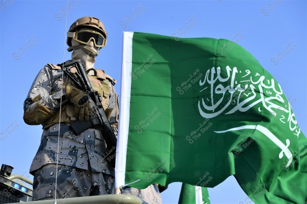 Image of a soldier in camouflage military uniform holding a rifle. The soldier stands next to a flag, which features the emblem of Saudi Arabia. The flag is green with the Islamic declaration of faith and a sword in the center. The soldier is wearing a helmet, goggles, and a tactical vest, with a clear blue sky in the background.