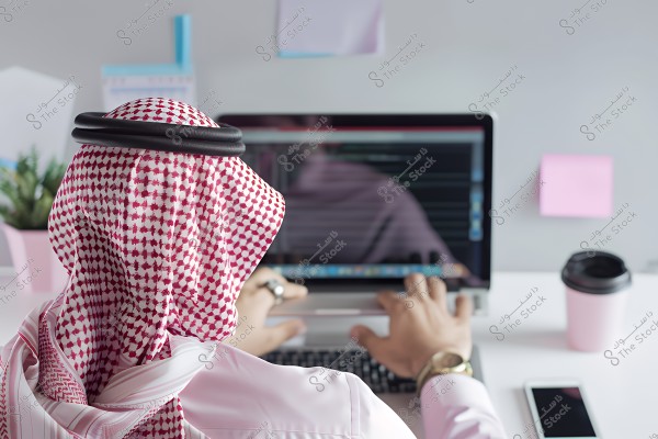 An image of a man sitting at a desk working on a laptop. He is wearing a red and white ghutra and agal, suggesting he might be from Saudi Arabia or a Gulf region. Only his hands are visible as he types on the keyboard. The desk also has a beverage cup, a smartphone, and a decorative plant in the background. There are also sticky notes on the wall.