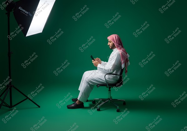 A man sits on a swivel office chair in a photography studio, wearing traditional Saudi attire including a thobe and a red and white checkered headscarf. He is looking at a mobile phone in his hand, with studio lighting positioned on the left. The background is green, complementing his clothing.