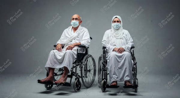 An elderly man and woman sitting in wheelchairs, wearing white Ihram clothing, with medical masks on their faces. Both are facing the camera in a studio setting with a uniform grey background.