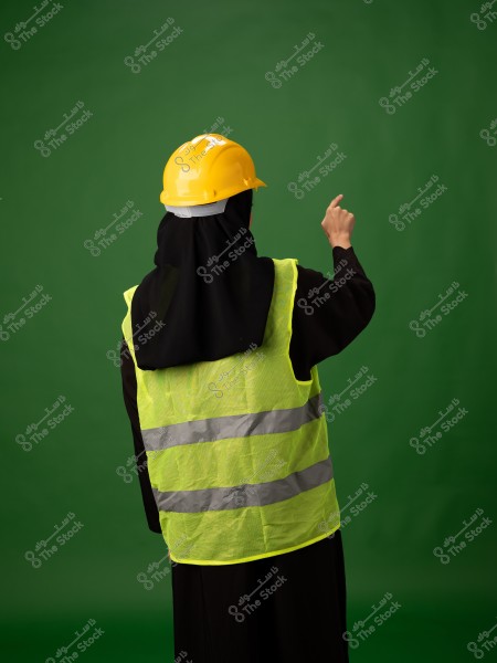 Image of a person wearing a yellow helmet and a yellow reflective safety vest over a black abaya, suggesting they may be from a Gulf region such as Saudi Arabia. The person stands against a green background, raising their right hand pointing upwards.