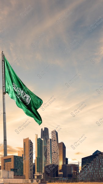 The image shows the green Saudi Arabian flag waving on a pole in the foreground against the skyline of Riyadh in Saudi Arabia. A collection of modern skyscrapers and contemporary architectural designs can be seen. The cloudy blue sky stretches above the scene, giving the image a sense of grandeur and urban development.