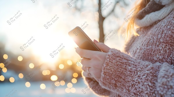 امرأة ترتدي سترة صوفية سميكة بألوان فاتحة تقف في الخارج وتحمل هاتفًا ذكيًا في يديها. يظهر في الخلفية السماء المشمسة مع تأثير البوكيه من الأضواء الدافئة البعيدة.