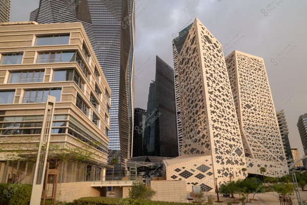 The image captures a cluster of modern skyscrapers in an urban area. On the left side, there is a building with a glass facade and horizontal lines, while in the center, there is a tower with a distinctive geometric design featuring square patterns. In the background, other towers with glass facades beautifully reflect the light. The sky is overcast, adding a dramatic touch to the scene. The foreground contains some small plants.